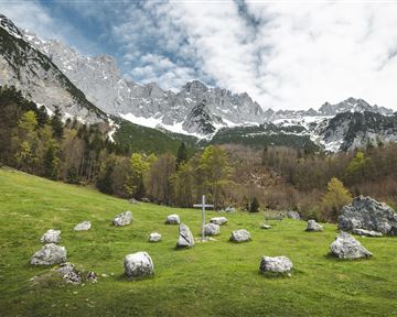 Eine grüne Wiese mit einer Steinkreisanlage und einem Holzkreuz im Vordergrund. Im Hintergrund erheben sich majestätische Berge unter einem wolkigen Himmel.