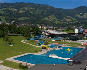 Ein großer Pool mit Rutschen und Liegewiesen in einer grünen Umgebung. Im Hintergrund sind Berge und ein klarer blauer Himmel zu sehen.