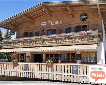 Ein schönes Holzgebäude mit einer einladenden Terrasse. Das Restaurant „Fabels“ hat ein rustikales Design und ist von Bäumen umgeben.