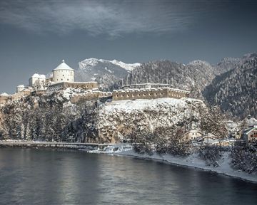 An impressive castle on a snowy hill with mountains in the background. The river reflects the winter landscape.