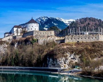Eine historische Burg auf einem Hügel mit schneebedeckten Bergen im Hintergrund. Ein klarer Fluss fließt im Vordergrund.