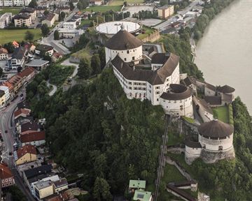Eine beeindruckende Burg auf einem Hügel mit umliegenden Häusern und einem Fluss. Die Landschaft ist grün und malerisch.