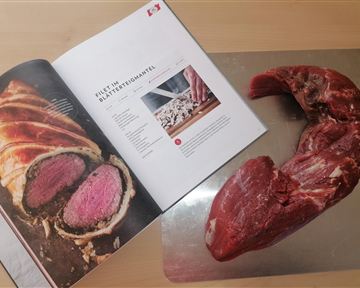Ein frisches Stück Fleisch liegt neben einem Kochbuch. Die Seite zeigt ein Rezept für ein Filetgericht.