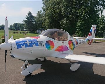 A colorfully decorated airplane is on a parking lot. In the background, trees and a green meadow can be seen.