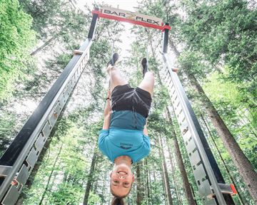 Ein junger Mann hängt kopfüber an einem Trainingsgerät im Wald. Die Umgebung ist grün und lichtdurchflutet.