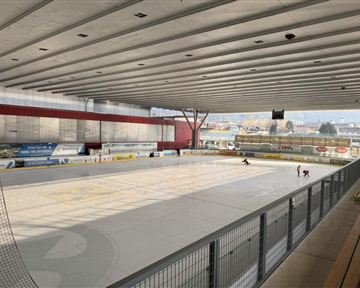 Eine Eishalle mit einer leeren Eisfläche und hohen Decken. Im Hintergrund sind einige Skater zu sehen.