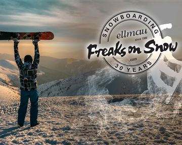 A snowboarder stands on a mountain peak holding his snowboard high. In the background, snowy mountains and a sunny sky can be seen.