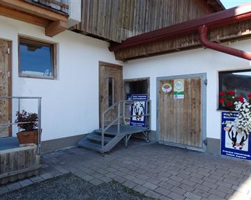 A building with wood siding and two doors. In front of the entrance stairs are plants and posters.