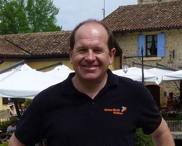 Ein Mann steht lächelnd vor einem historischen Gebäude. Im Hintergrund sind Bäume und Tische im Freien zu sehen.