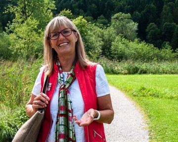 Eine freundliche Frau lächelt auf einem Wanderweg umgeben von grüner Natur. Im Hintergrund sind Bäume und eine sonnige Landschaft zu sehen.