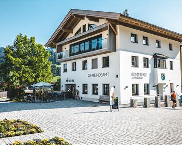 Ein modernes Gebäude mit der Aufschrift "Gemeindeamt". Vor dem Gebäude sind Bäume, Blumenbeete und einige Menschen zu sehen.