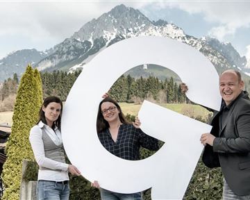 Drei Personen halten einen großen Buchstaben "G" vor einer beeindruckenden Berglandschaft. Im Hintergrund sind schneebedeckte Berge und grüne Bäume sichtbar.