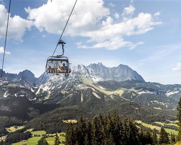 A cable car travels over a dreamy mountain landscape. In the background, tall mountains and green meadows can be seen under a clear sky.