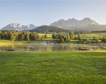 A picturesque landscape with a tranquil lake and gentle hills. In the background, majestic mountains rise under a clear sky.