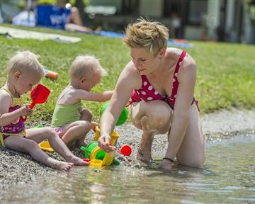 Eine Mutter spielt mit ihren zwei kleinen Kindern am Ufer. Sie haben Spielzeug und genießen das Wasser an einem sonnigen Tag.