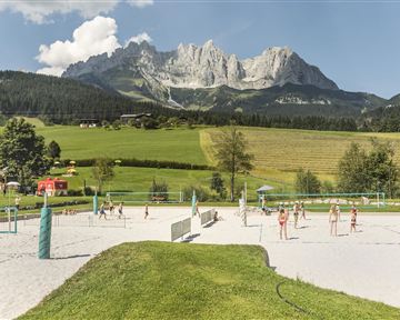 A sandy court with volleyball players in front of impressive mountains. The surroundings are green and sunny, ideal for sports activities.