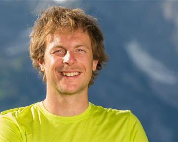 A smiling man is wearing a bright green shirt. Blurred mountains are visible in the background.