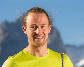 Ein lächelnder Mann in einem leuchtend gelben Shirt. Im Hintergrund sind Berge mit schneebedeckten Gipfeln zu sehen.