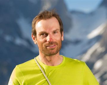A man stands in front of a mountainous backdrop. He is wearing a light green sports shirt and has a friendly expression.