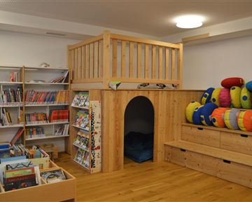 Ein einladender Raum mit einem Lesebereich, Bücherregalen und einem gemütlichen Rückzugsort. Bunte Kissen bieten Platz zum Sitzen und Entspannen.