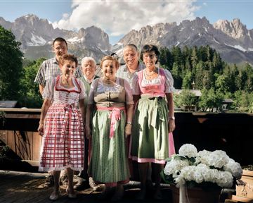 A group of six people in traditional costumes stands in front of an impressive mountain landscape. In the background, green forests and high mountains are visible.