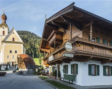Ein traditionelles altes Gasthaus mit Holzverzierung und grünen Fensterläden steht neben einer schönen Kirche. Im Hintergrund sind grüne Hügel und eine klare Himmel sichtbar.