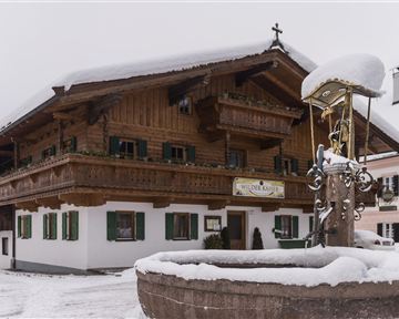 A traditional wooden house in the snow with green shutters. In the foreground, there is a fountain surrounded by a wintry landscape.