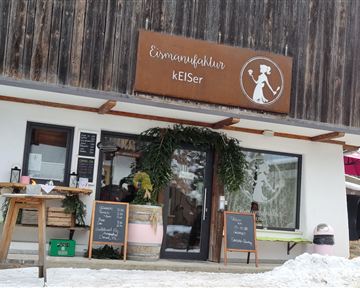 A cozy café with a rustic wooden sign. Outside, there is snow and decorative plants.