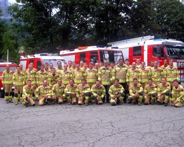 A group of firefighters stands in striking uniforms in front of several fire trucks. They pose with serious expressions on a paved area.