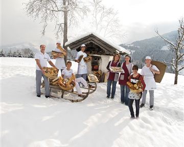 Eine Gruppe von Menschen steht im Schnee vor einer kleinen Hütte. Sie halten frisches Brot und Gebäck in den Händen.