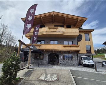 A modern building with wooden cladding and large windows. In front of it, there are flags and a parking lot is available.