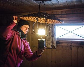 A person stands in a wooden room and holds a lamp over a basket. It is dark, but the light from the lamp illuminates the room.