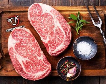 Zwei große Stücke hochwertige Rindfleisch auf einem Holzbrett, umgeben von Gewürzen und frischen Kräutern. Daneben liegt ein Schaufelmesser und Salz in einer kleinen Schale.