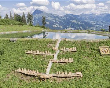 Eine malerische Landschaft mit einer Holzliegefläche inmitten von grünen Wiesen und einem ruhigen Teich. Im Hintergrund sind majestätische Berge und ein blauer Himmel zu sehen.