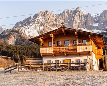 Ein traditionelles Holzhaus in den Bergen, umgeben von schneebedeckten Gipfeln. Die Landschaft ist friedlich und idyllisch.