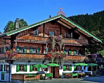 A traditional alpine house made of wood with blooming plants and green windows. In front of the building, there are seating areas with umbrellas.