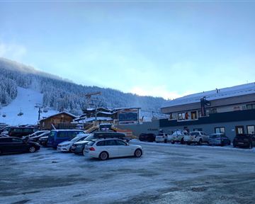Ein Parkplatz in den Bergen, umgeben von schneebedeckten Hügeln. Im Hintergrund sind Skilifte und Gebäude zu sehen.