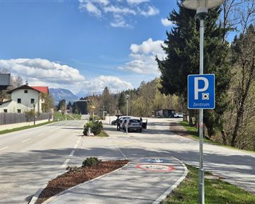 Eine ruhige Straße mit Parkplätzen und einigen Häusern auf der linken Seite. Im Hintergrund sind Bäume und Berge zu sehen, und der Himmel ist klar und blau.