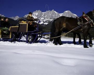 Eine Kutsche mit Pferd fährt durch den Schnee, während Menschen darin sitzen. Im Hintergrund sind beeindruckende Berge unter einem klaren blauen Himmel zu sehen.