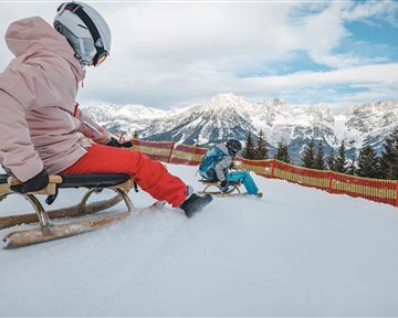 Two children are sledding down a snowy slope. In the background, snow-covered mountains and a clear sky can be seen.