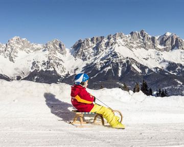 A child is sitting on a sled in the snow. In the background, majestic mountains and a clear blue sky can be seen.