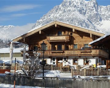 A traditional, wooden-clad chalet in a snowy mountain landscape. In the background, impressive mountains and a clear blue sky can be seen.