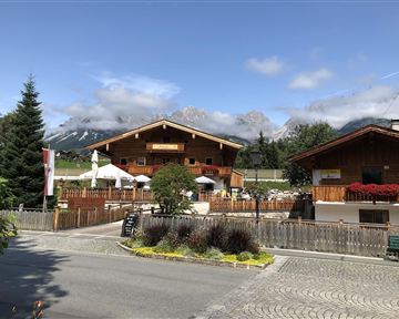 An idyllic chalet in the mountains with cozy terraces. The surroundings are green with a view of high mountains and a clear sky.