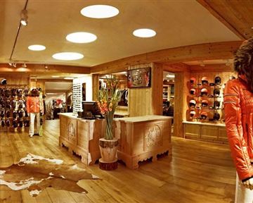 A modern sports shop with paneled wooden walls and warm lighting. In the foreground is a stylish mannequin in an orange jacket, surrounded by ski equipment and accessories.