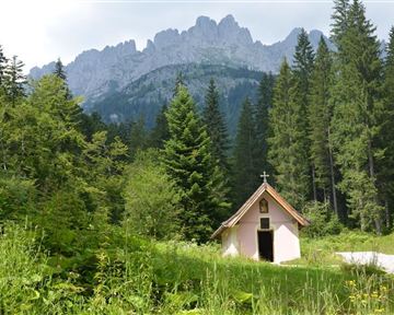 A small chapel stands at the edge of a forest, surrounded by tall trees. Majestic mountains rise in the background.