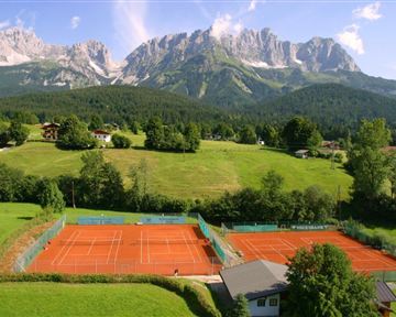 Two tennis courts on red clay, surrounded by lush greenery and mountains in the background. The sky is clear and sunny.