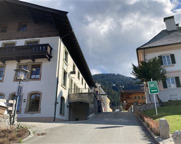 A charming street in the mountains with traditional buildings. In the background, the green hills are visible along with a sign for a restroom.