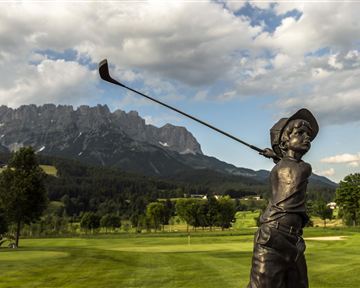 A bronze sculpture of a young golfer stands on a golf course. In the background, majestic mountains and a clear sky can be seen.