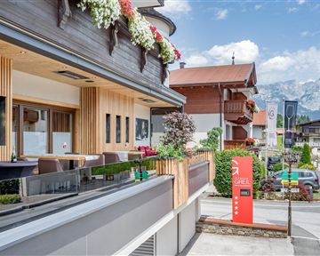 Eine moderne Fassade mit Holzverkleidung und blühenden Blumen. Im Hintergrund sind Berge und ein klarer Himmel zu sehen.