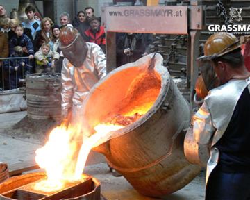 Zwei Personen in Schutzkleidung gießen flüssiges Metall aus einem großen Kessel. Im Hintergrund stehen Zuschauer, die das Spektakel beobachten.
